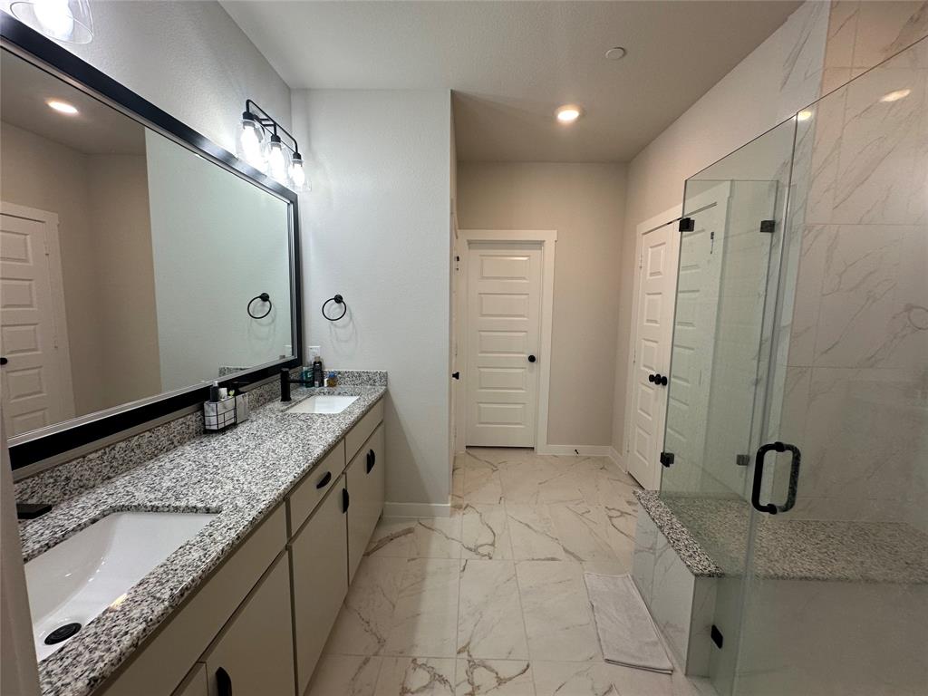 Spa-inspired primary bathroom featuring dual vanities with speckled quartz countertops, a frameless glass-enclosed shower with marble-look tile surround, and recessed ceiling lighting Spa-inspired primary bathroom featuring dual vanities with speckled quartz countertops, a frameless glass-enclosed shower with marble-look tile surround, and recessed ceiling lighting