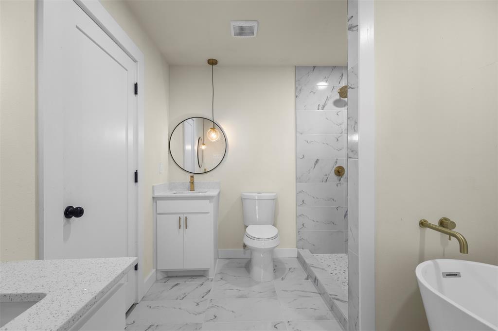 White vanity cabinet with quartz countertop, circular wall-mounted mirror, brass-finish fixtures, stone-patterned tile flooring, and a walk-in shower with stone-patterned tile surround