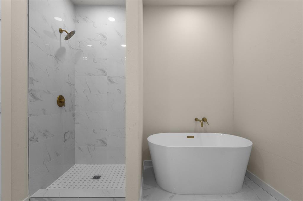 Freestanding soaking tub, walk-in shower with glass enclosure, white marble-look tile walls, and gold-finish fixtures
