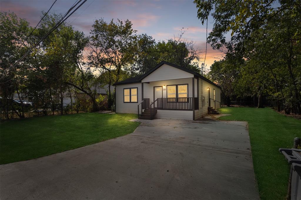 White exterior siding with dark trim, covered front porch, concrete driveway, manicured lawn, and mature trees White exterior siding with dark trim, covered front porch, concrete driveway, manicured lawn, and mature trees