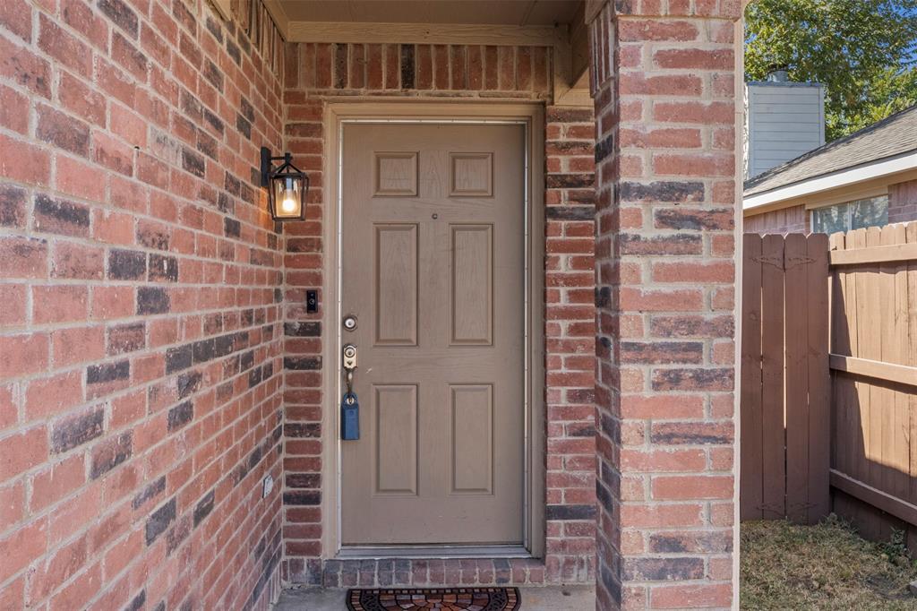 Brick exterior facade with a paneled entry door, exterior wall lantern, and partial wood privacy fence Brick exterior facade with a paneled entry door, exterior wall lantern, and partial wood privacy fence