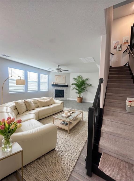 Open concept living area featuring wood-finish flooring, multiple windows with blinds, a modern ceiling fan, and a fireplace with a dark mantel Open concept living area featuring wood-finish flooring, multiple windows with blinds, a modern ceiling fan, and a fireplace with a dark mantel