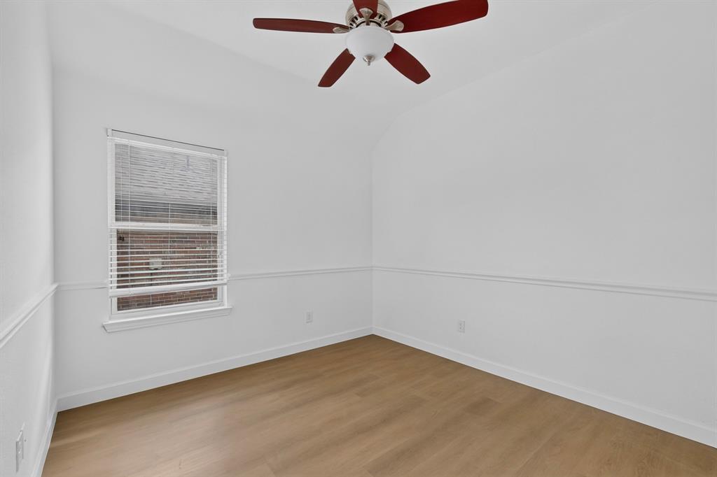 Neutral-toned interior space featuring wood-finish flooring, white wall molding, a single-hung window with blinds, and a multi-blade ceiling fan with integrated lighting