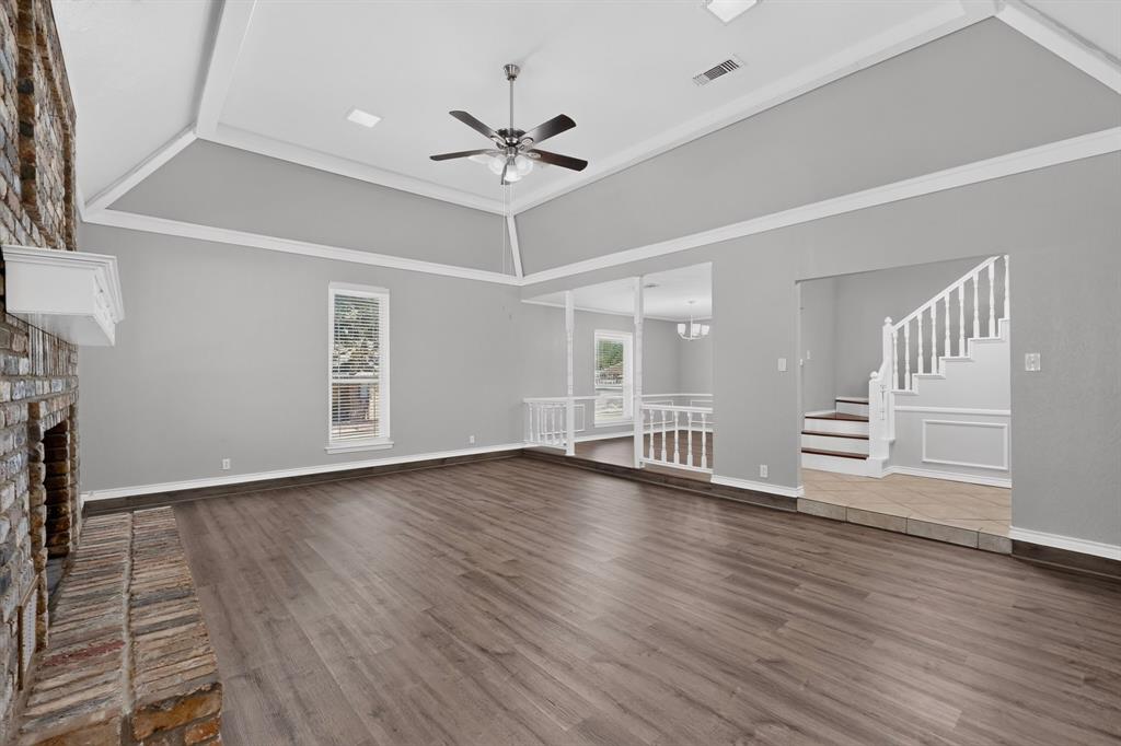 Spacious room featuring vaulted ceilings, a prominent brick fireplace with a white mantel, wood-finish flooring, and a staircase with white balusters