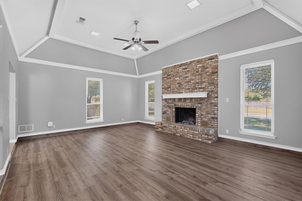 Spacious room featuring a vaulted ceiling, wood-finish flooring, a brick fireplace with a white mantel, and multiple windows with blinds