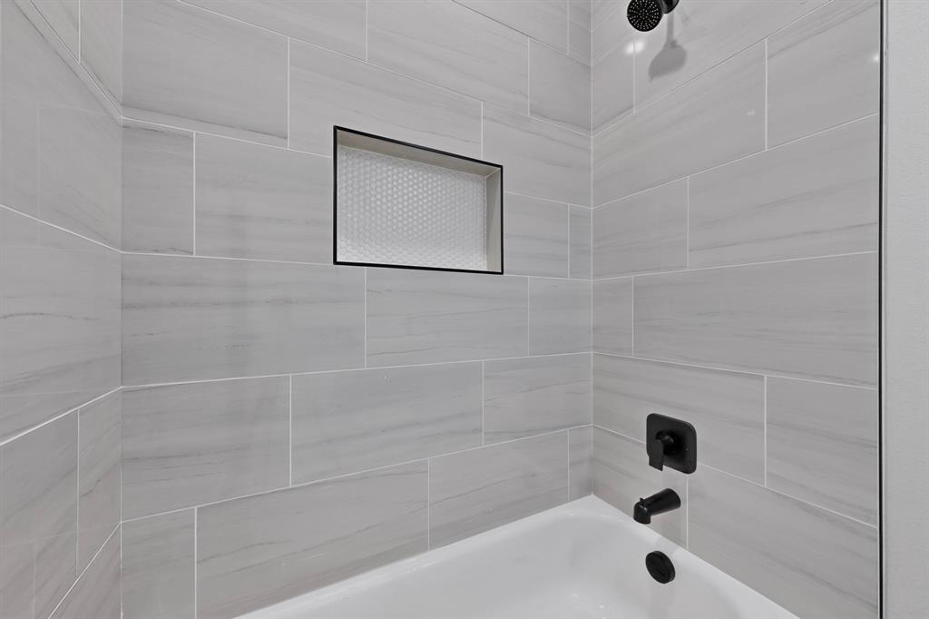 Bathtub with matte black fixtures, an integrated shower niche with honeycomb tile, and light gray subway tile surround