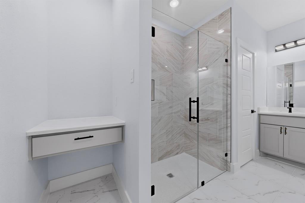 Corner vanity with drawer, glass-enclosed shower with tile surround, single vanity with under-sink storage, and large format floor tiling