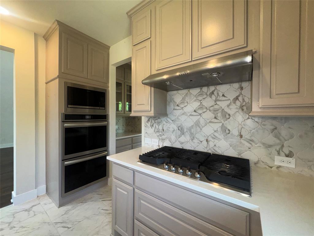 Light-toned cabinetry with crown molding, double wall ovens, integrated microwave, stainless steel range hood, and a decorative tile backsplash