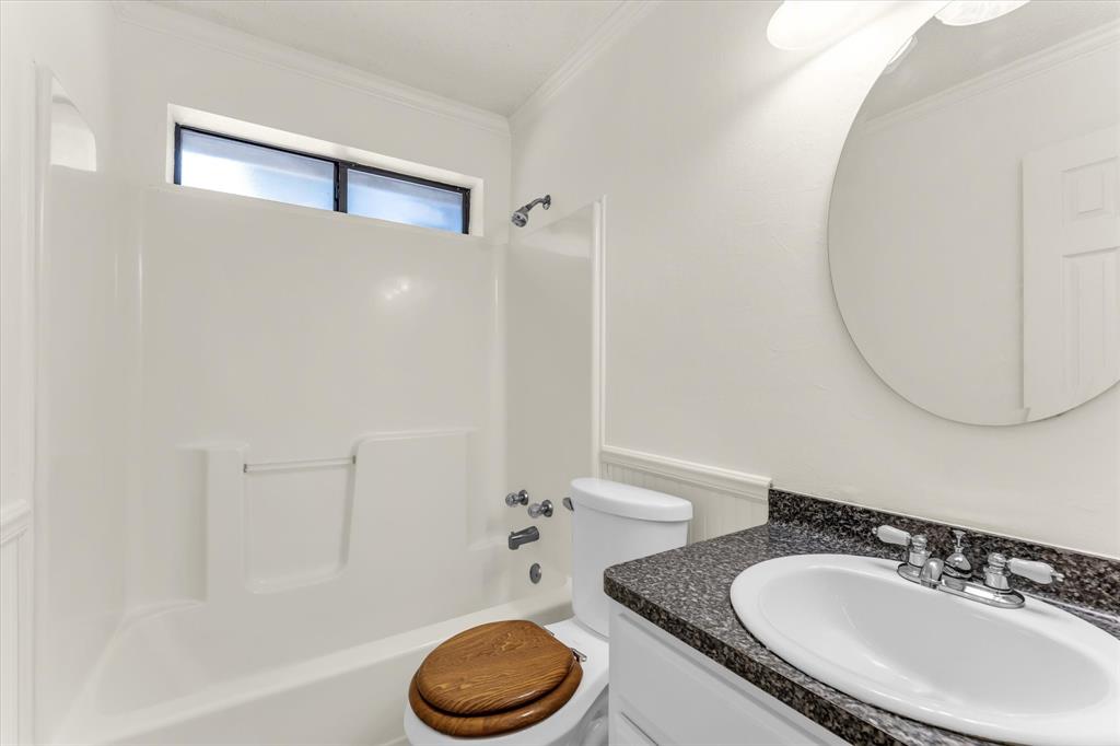 White bathtub and shower combination with chrome fixtures, transom window, white vanity with dark speckled countertop, and a circular wall mirror