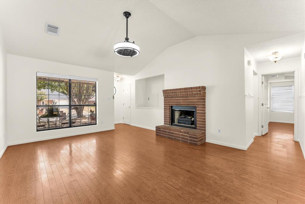 Vaulted ceiling, wood-finish flooring, brick fireplace, large window, and ceiling-mounted light fixture