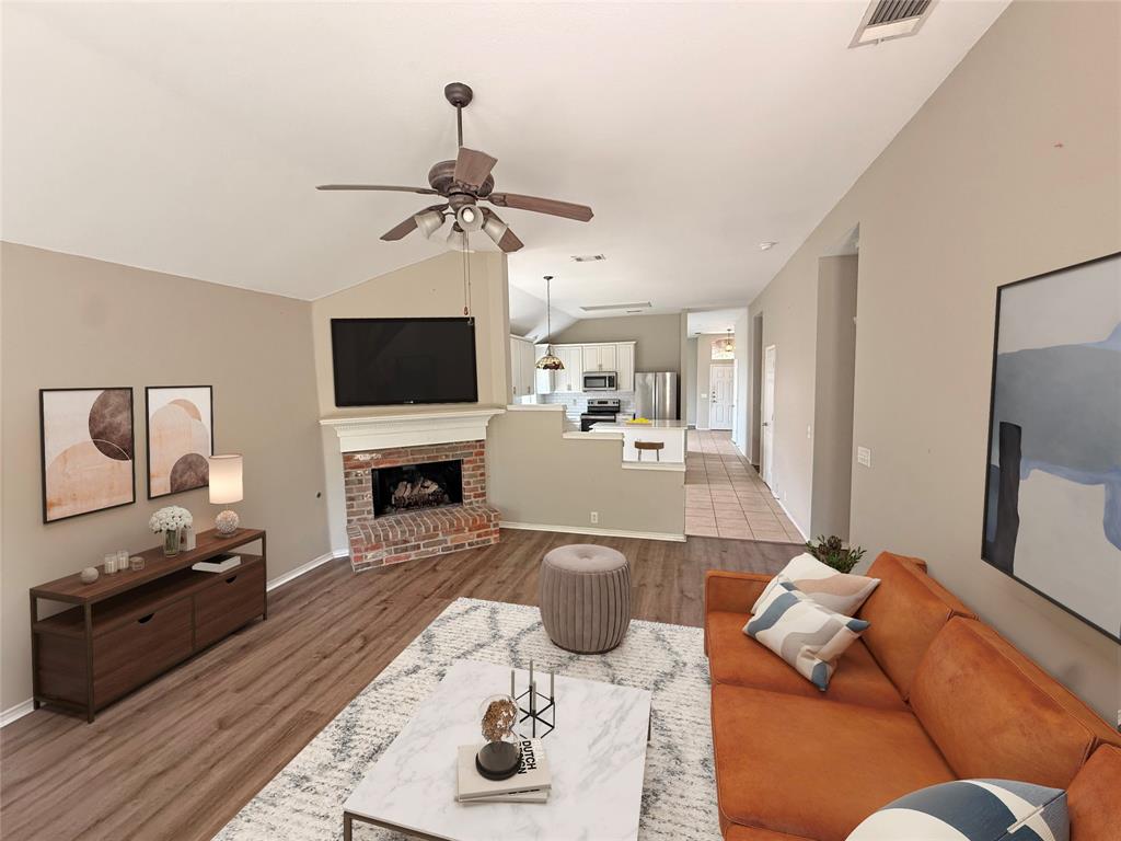 Vaulted ceiling with a ceiling fan, wood-finish flooring, brick-faced fireplace with a white mantel, kitchen with white cabinetry, and tile flooring