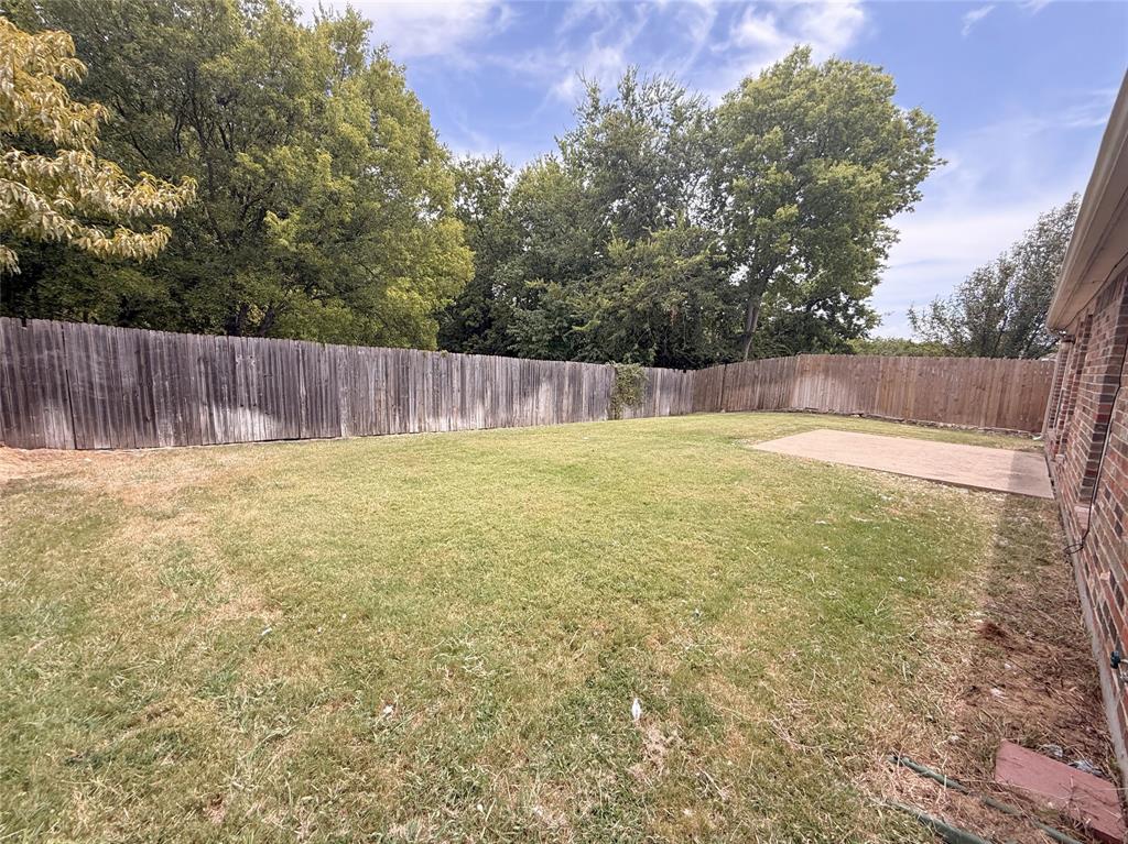 Expansive backyard featuring a wood fence, mature trees, a concrete patio, and a brick exterior wall
