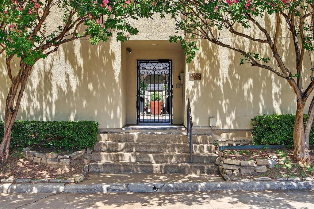 Private guest parking behind building. Stucco exterior with mature trees, stone-edged landscaping, and a wrought iron entry gate.