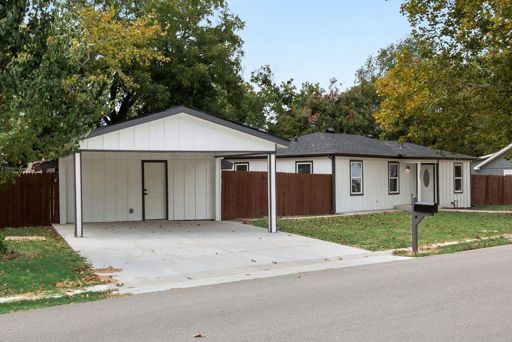 White siding exterior with dark trim accents, a covered carport, dark shingle roof, and a wood fence enclosing the property