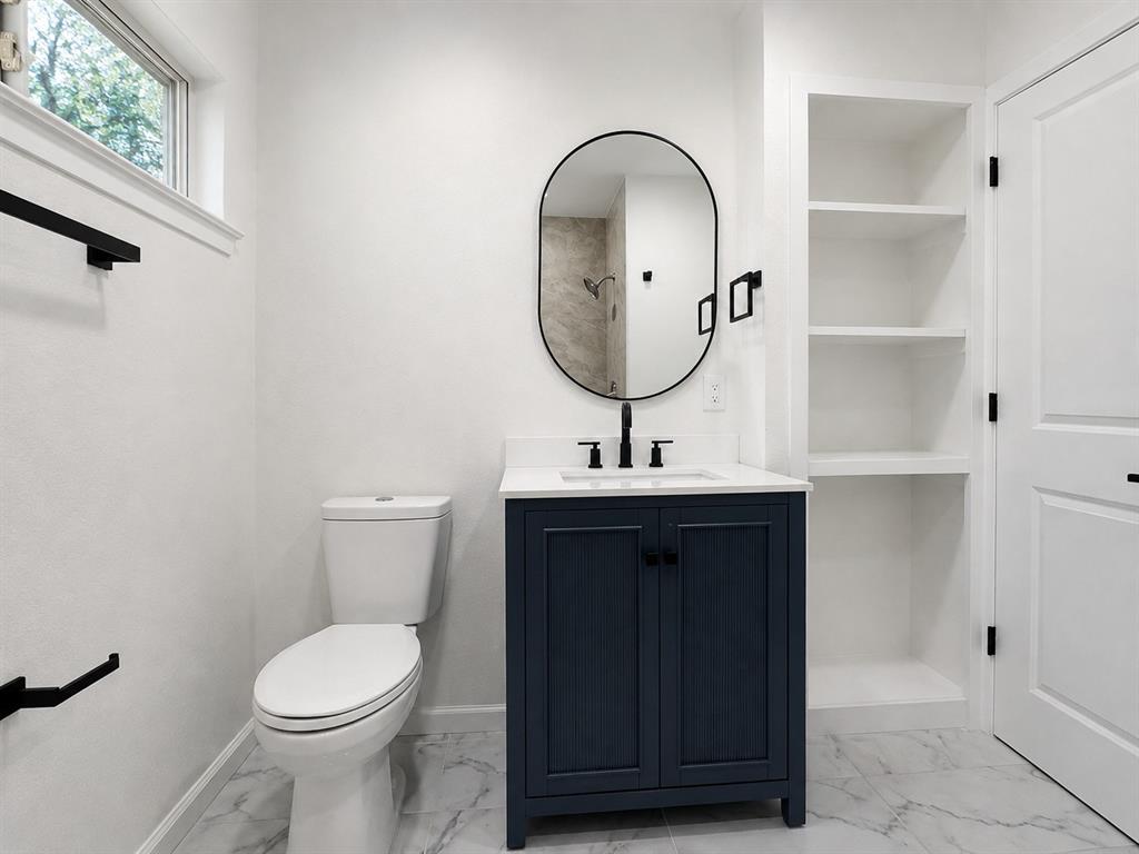 White marble-look flooring, dark blue vanity with a white countertop and undermount sink, built-in shelving, and a black-framed oval mirror