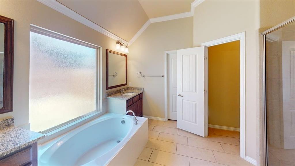 Soaking tub with a large privacy window, dual vanity sinks with stone countertops, and tile flooring