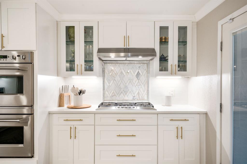 White shaker-style cabinetry with gold-tone hardware, stainless steel double ovens, gas cooktop, glass-front upper cabinets, and a chevron-patterned tile backsplash