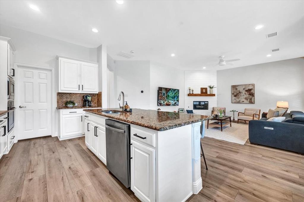 Wood-finish flooring throughout, kitchen island with granite countertop and integrated sink, stainless steel dishwasher, white cabinetry with black hardware, and a living area with a fireplace featuring a shiplap accent wall