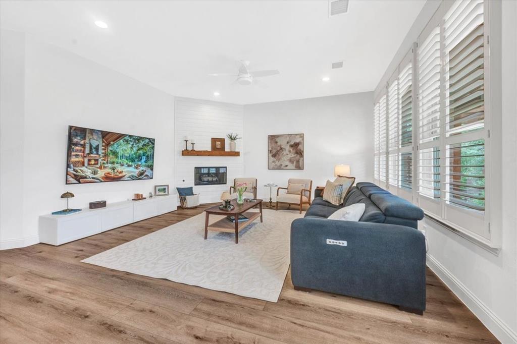Spacious living area featuring wood-finish flooring, a white shiplap fireplace with a wood mantle, recessed lighting, and multiple windows with plantation shutters