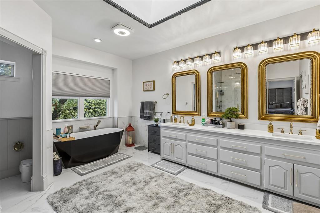 Freestanding soaking tub, dual vanity with two sinks, gold-framed mirrors, vanity lighting, and white tile flooring