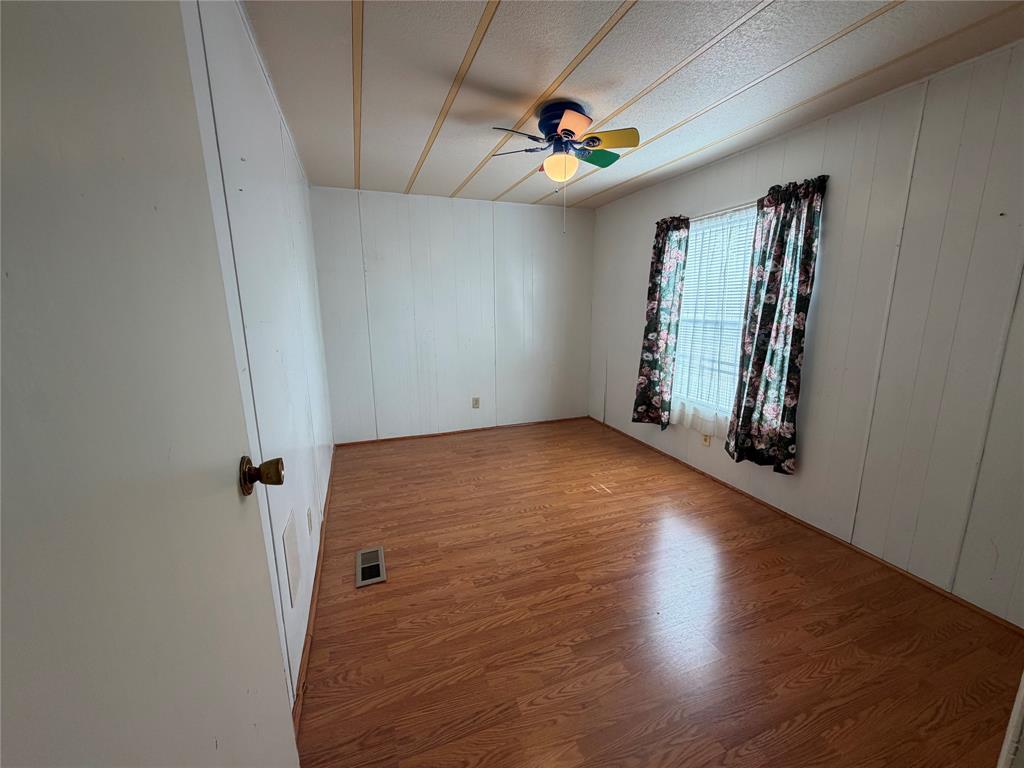 Spacious bedroom with warm wood-finish flooring, white wall paneling, ceiling-mounted fan with lighting fixture, and large window with patterned curtains allowing natural light
