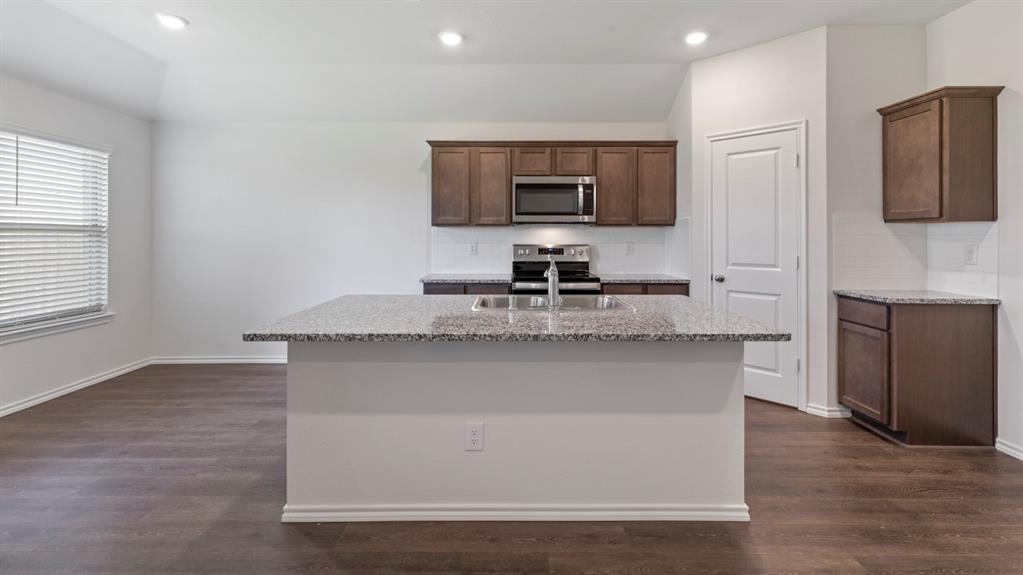 Modern kitchen featuring a large center island with granite countertop, stainless steel appliances including dual ovens, rich wood cabinetry with upper and lower storage, recessed lighting, and gleaming wood-finish flooring throughout the open-concept space