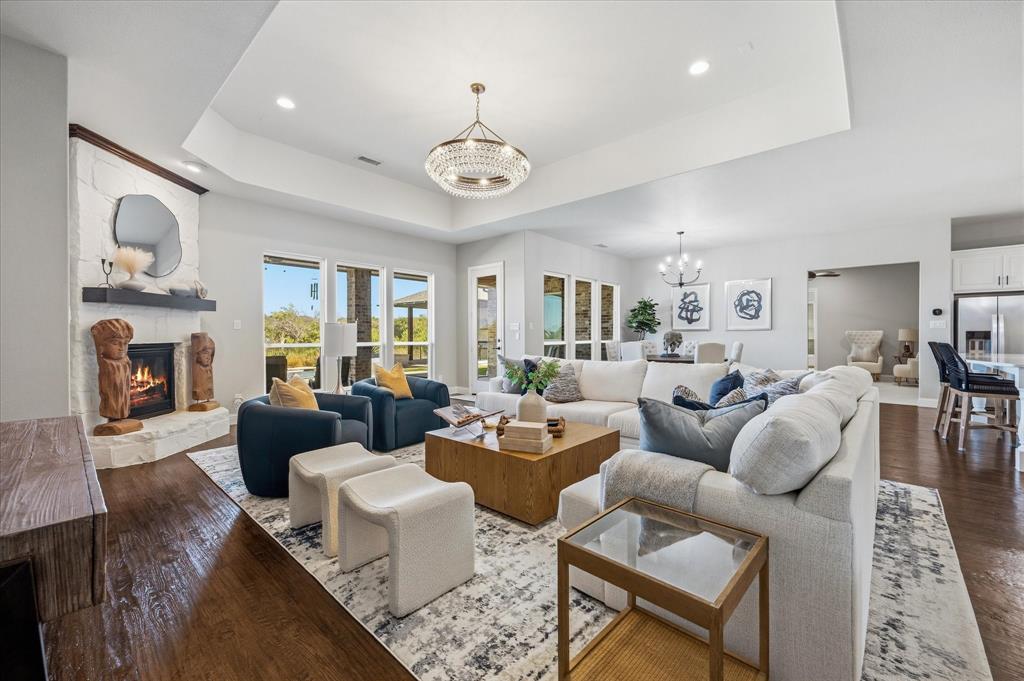 Expansive living area featuring wood-finish flooring, a white brick fireplace with a dark mantel, a tray ceiling, and large windows