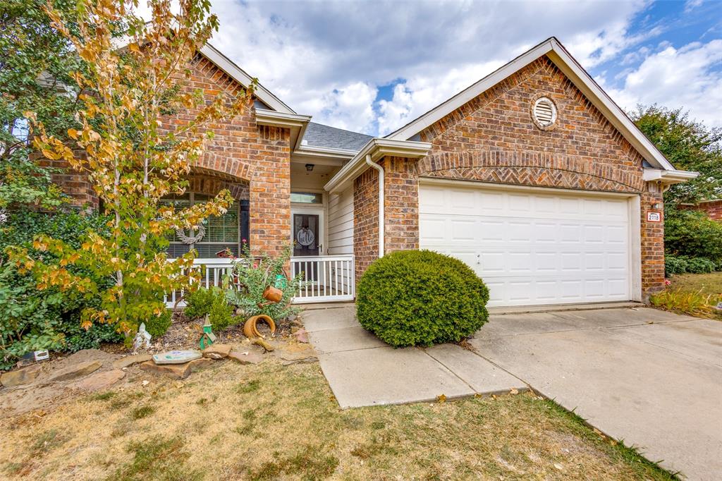 Charming brick home featuring a covered front porch with white railings, two-car garage with white paneled door, manicured landscape with mature shrubs, concrete driveway, and gable roofline with architectural detailing