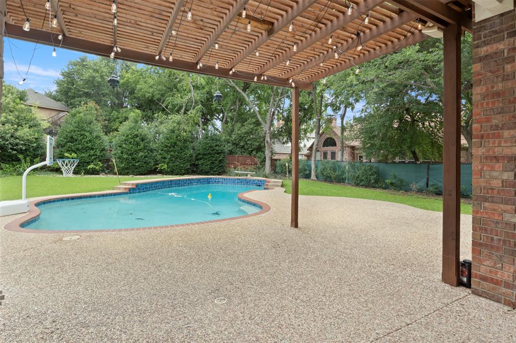 Expansive exposed aggregate patio with an attached wood pergola, complemented by a curved in-ground swimming pool featuring blue tile accents and an adjacent green lawn
