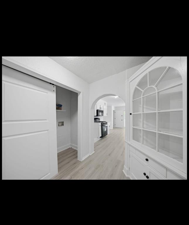 Light wood-finish flooring, white barn door, built-in display cabinet with glass doors, arched doorway, and black kitchen appliances