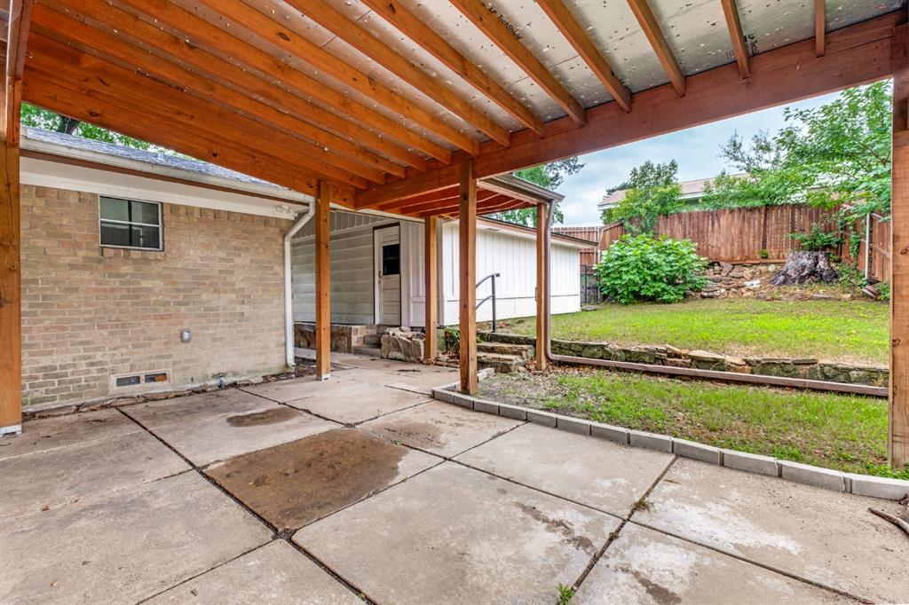 Covered patio with exposed wood beam ceiling and brick exterior wall, featuring concrete flooring and direct access to a spacious fenced yard with mature landscaping and wooden fence perimeter