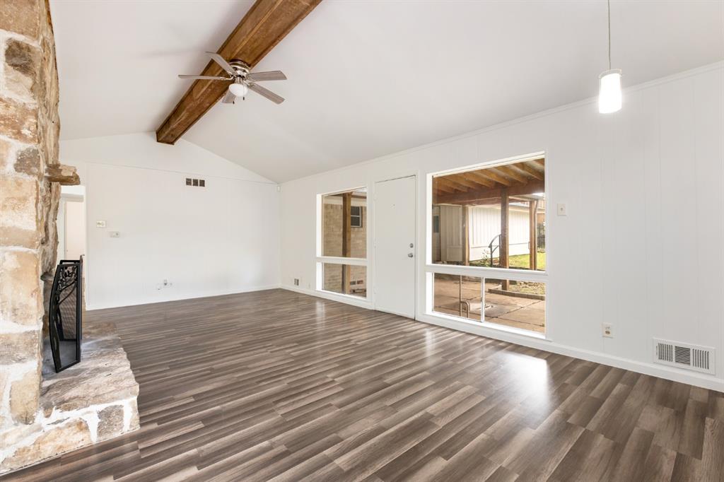 Vaulted ceiling with exposed wood beam, ceiling fan, wood-finish flooring, stone-clad fireplace, and large window