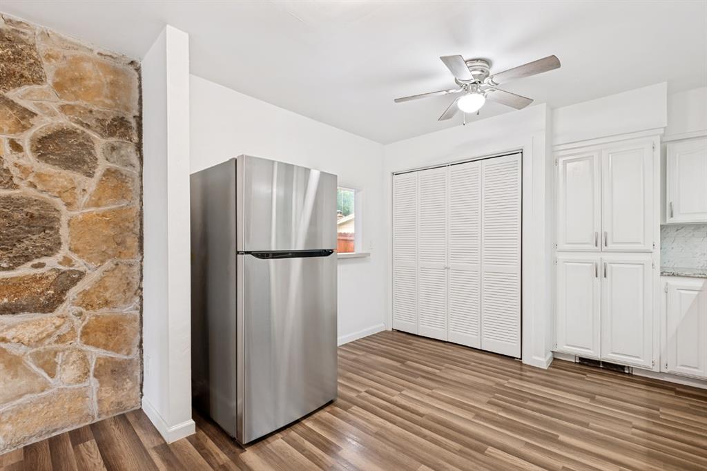Stone accent wall, wood-finish flooring, stainless steel refrigerator, white shaker-style cabinetry, and ceiling fan with integrated lighting