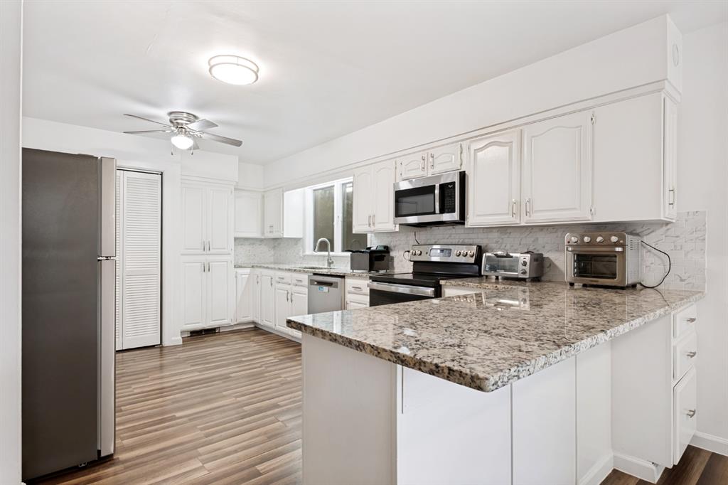 Modern kitchen featuring white cabinetry with ample storage, granite-style countertops with waterfall island edge, stainless steel appliances including gas range and microwave, and wood-finish flooring throughout the open layout