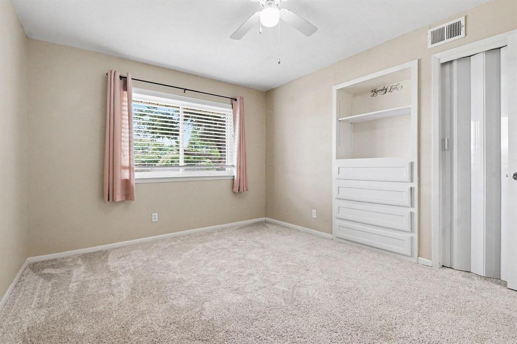 Light-colored carpeted flooring, built-in shelving with integrated drawers, ceiling fan with light fixture, and horizontal window blinds
