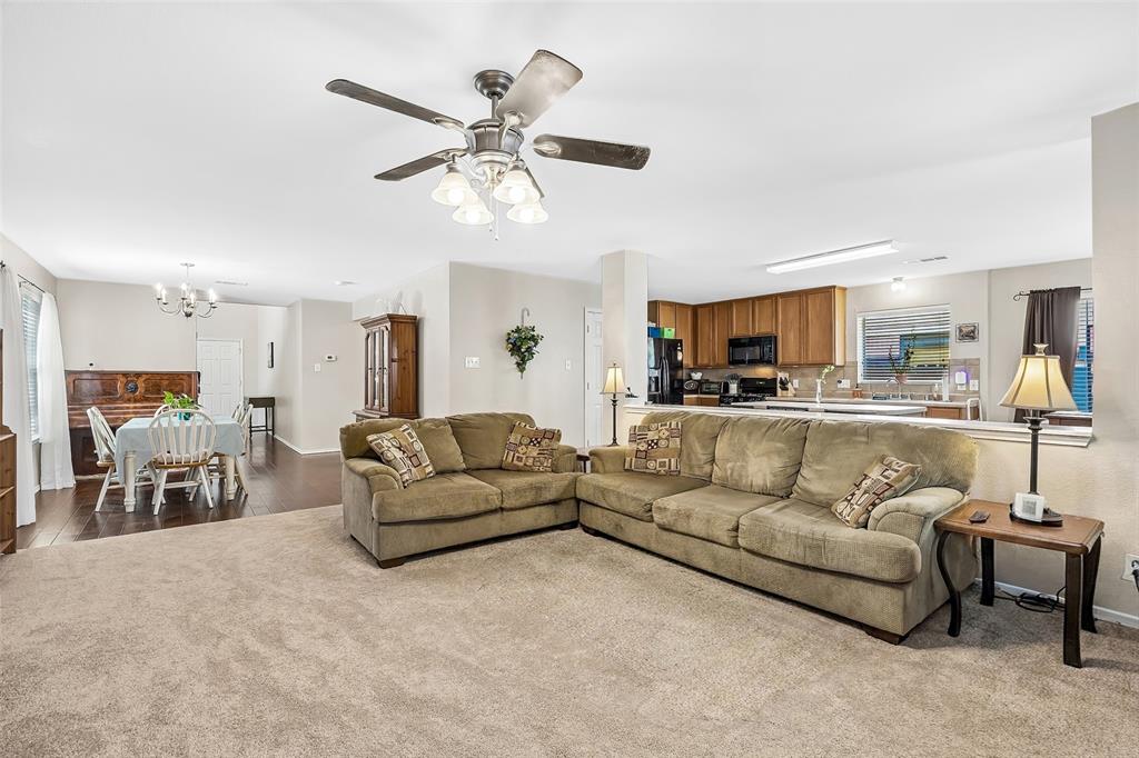 Expansive living area with neutral carpeting, an overhead ceiling fan with integrated lighting, and direct access to a kitchen featuring wood cabinetry and black appliances