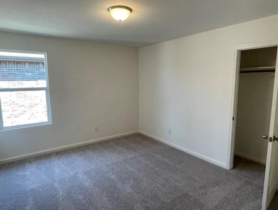Wall-to-wall carpeting in a neutral tone, ceiling-mounted light fixture, white interior walls, and a window with a brick exterior visible