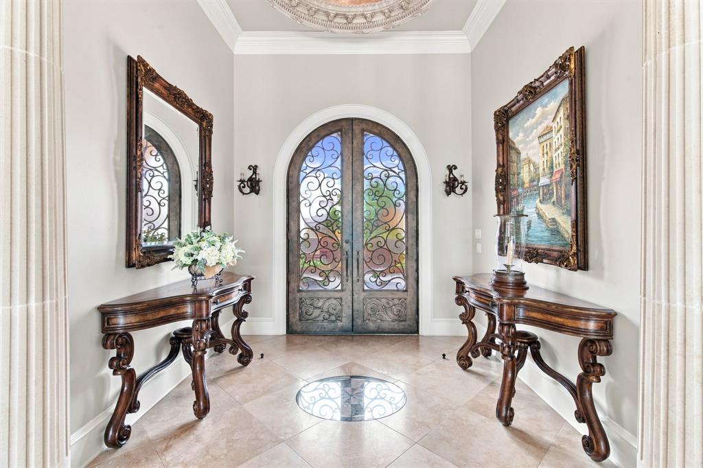 Arched entryway with ornate ironwork double doors, tile flooring with a decorative inlay, crown molding, and fluted architectural columns