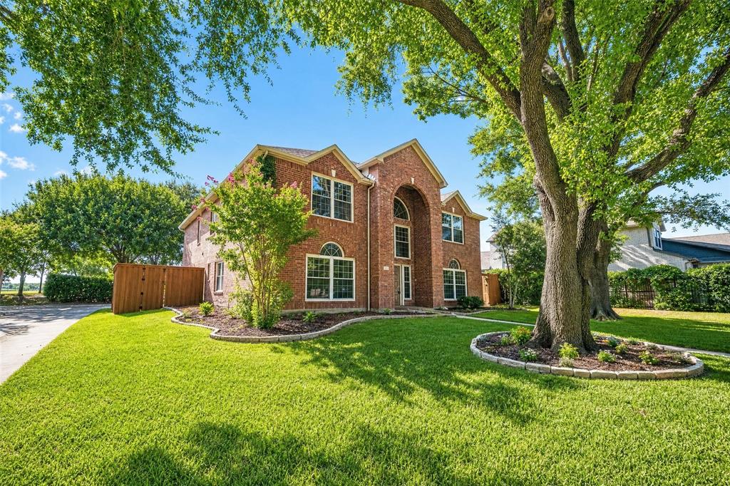 Traditional brick exterior with arched entry, lush green lawn, mature trees, and landscaped beds with stone edging
