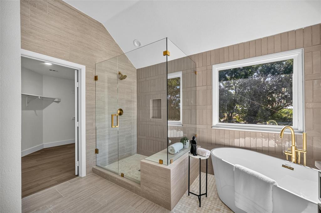 Free-standing soaking tub with brass fixtures, glass-enclosed shower with brass hardware, tiled shower niche, wood-finish flooring, and a walk-in closet