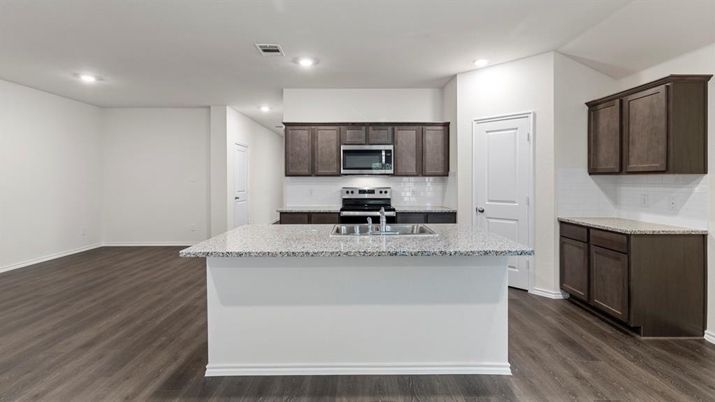 Kitchen featuring a center island with a built-in sink, stainless steel appliances, dark wood-finish cabinetry, light-colored countertops, and white subway tile backsplash
