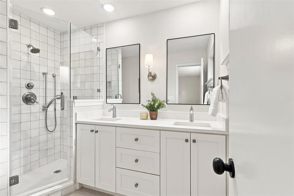 Glass-enclosed shower with subway tile surround, dual vanity with white cabinetry, quartz-finish countertops, and dual framed mirrors