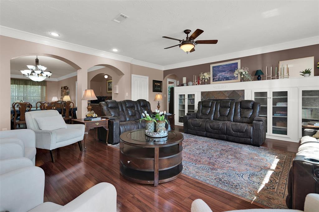 Hardwood-finish flooring throughout, tray ceiling with a built-in fan, custom built-in cabinetry with glass-front doors, decorative archways, and crown molding