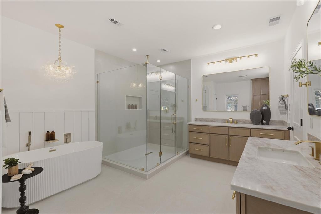 Freestanding soaking tub with vertical fluting, a spacious glass-enclosed shower, dual vanity with undermount sinks, stone countertops, and gold-toned hardware