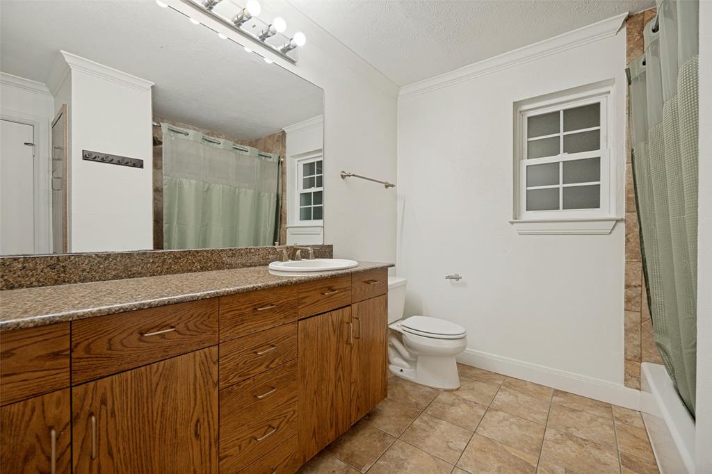 Wood-finish vanity with an integrated sink, granite-style countertop, large wall-mounted mirror, ceramic tile flooring, and a window providing natural illumination