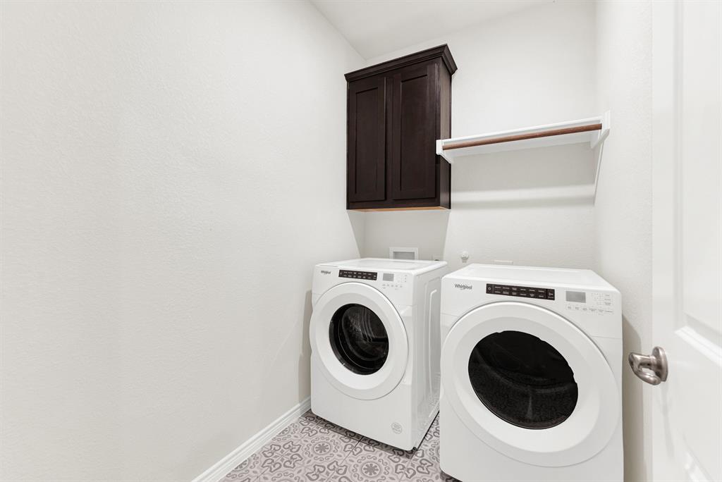 The functional utility room features patterned tile floors, upper cabinetry, and a dedicated hanging rod above modern front-load laundry units. Keep daily chores organized and hassle-free in this clean, stylish, and practical space.