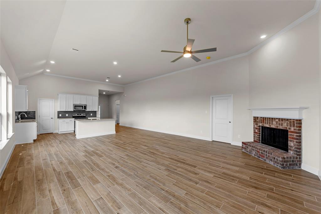 Spacious open-concept interior featuring vaulted ceilings, wood-finish flooring, a brick fireplace, and a kitchen with white cabinetry and stainless steel appliances