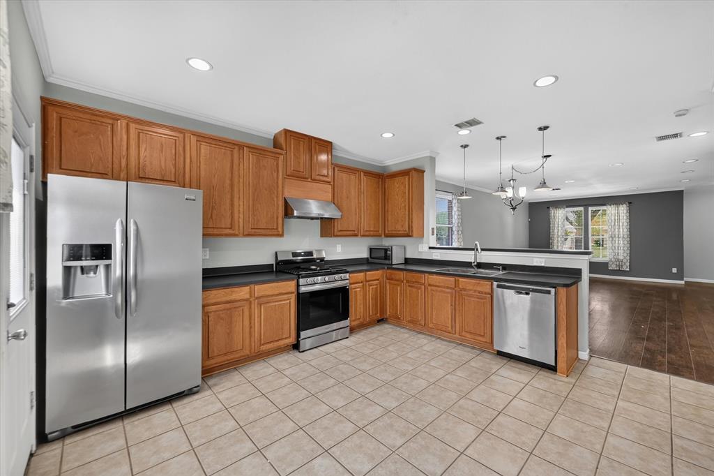 Spacious kitchen featuring warm wood cabinetry, stainless steel appliances including a side-by-side refrigerator and gas range, black countertops, tile flooring, recessed and pendant lighting, and a large island with seating area