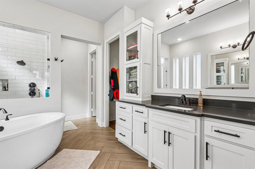 Freestanding soaking tub, walk-in shower with subway tile, dual vanity with undermount sink, dark countertop, and herringbone wood-finish flooring