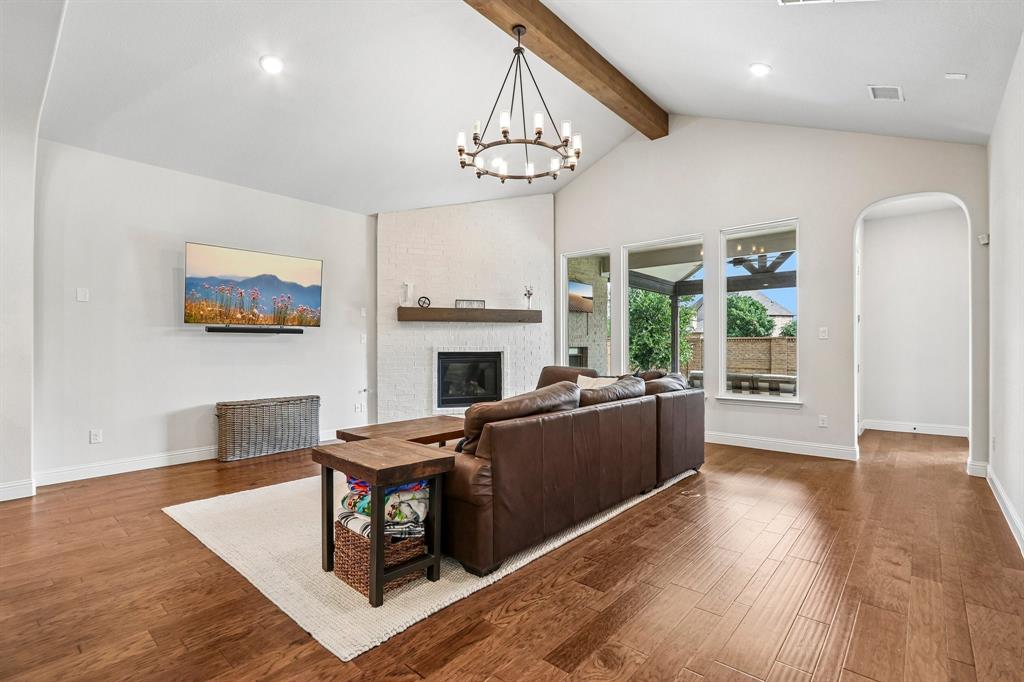 Vaulted ceiling with exposed wood beam, wood-finish flooring, white brick fireplace, arched doorway, and large windows